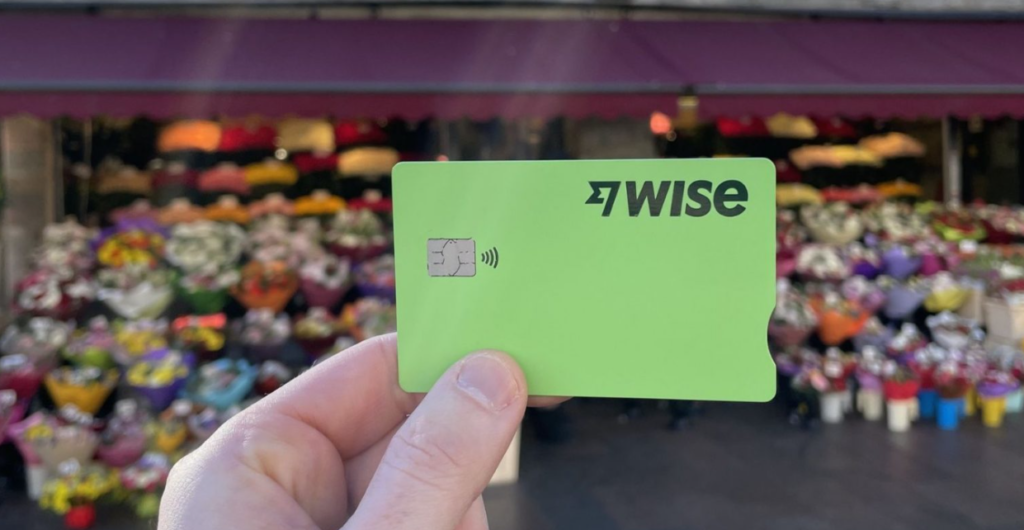 Mão segurando um cartão verde da Wise em frente a uma banca colorida, durante uma viagem.