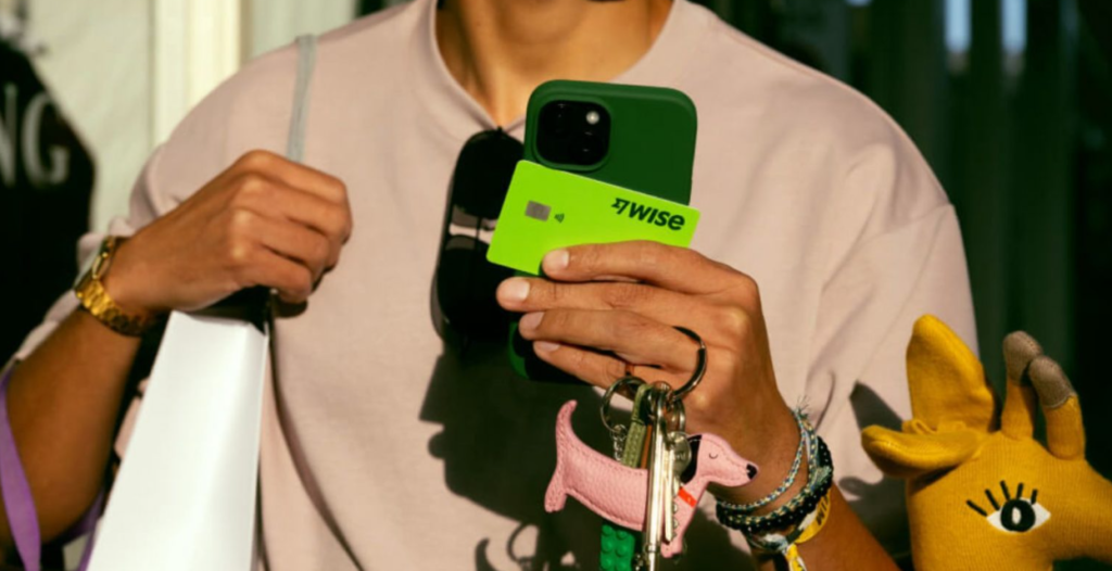Pessoa segurando um celular com um cartão verde da Wise, enquanto carrega uma bolsa e segura chaves, em uma cena relacionada a viagem.