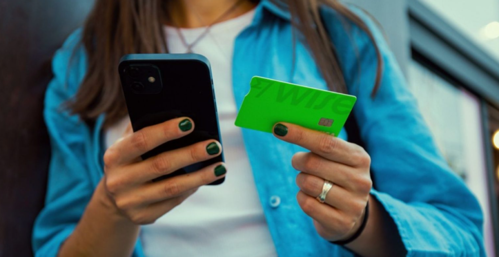 Mulher segurando um celular em uma mão e um cartão bancário verde na outra.