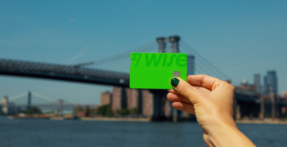 Mão segurando um cartão verde da Wise em frente à Ponte do Brooklyn, em Nova York, com o rio e o horizonte da cidade ao fundo.